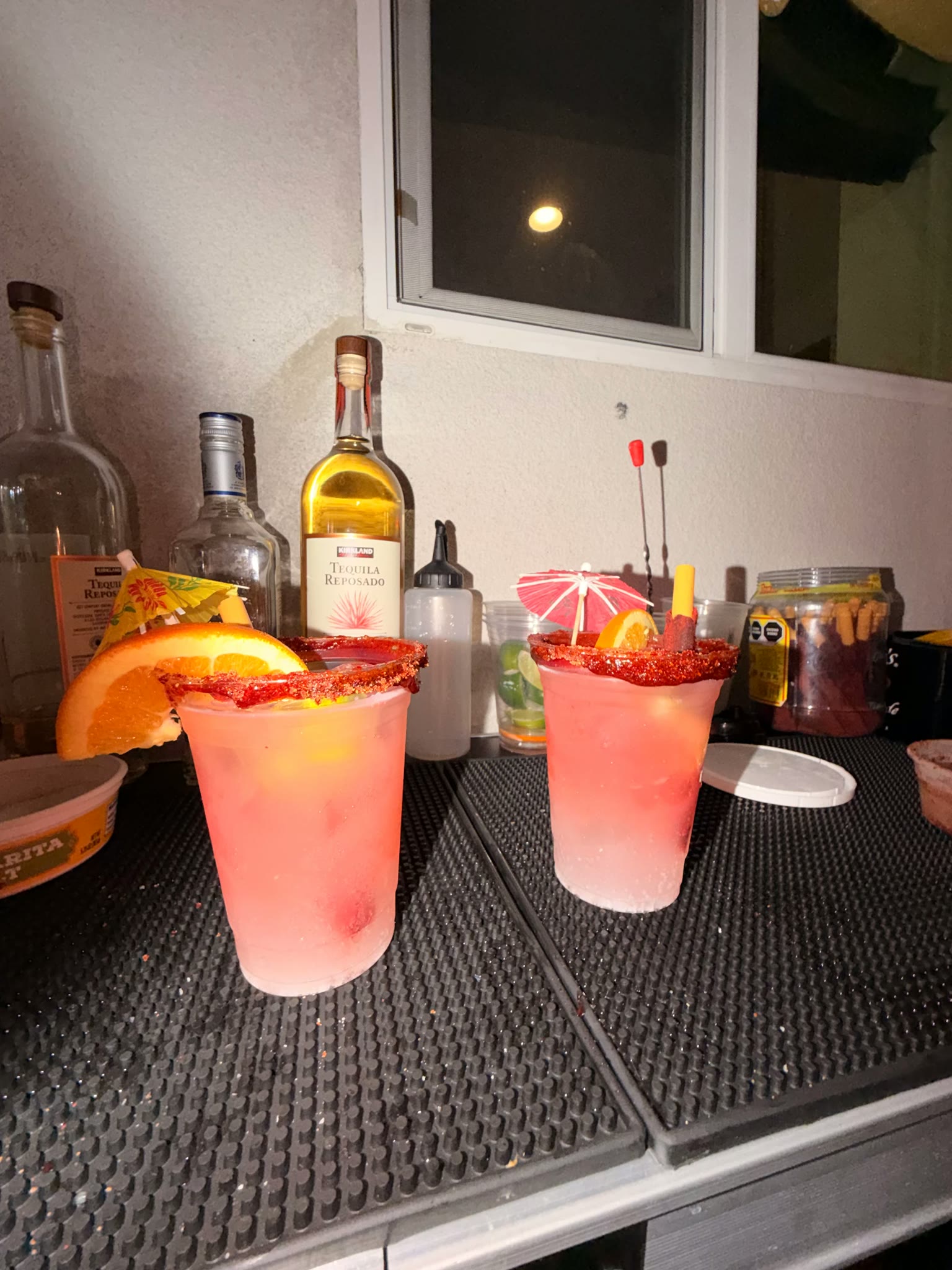 Two pink cocktails in clear cups with orange wedges, cocktail umbrellas and chili-salted rims, set on a black bar mat in front of liquor bottles